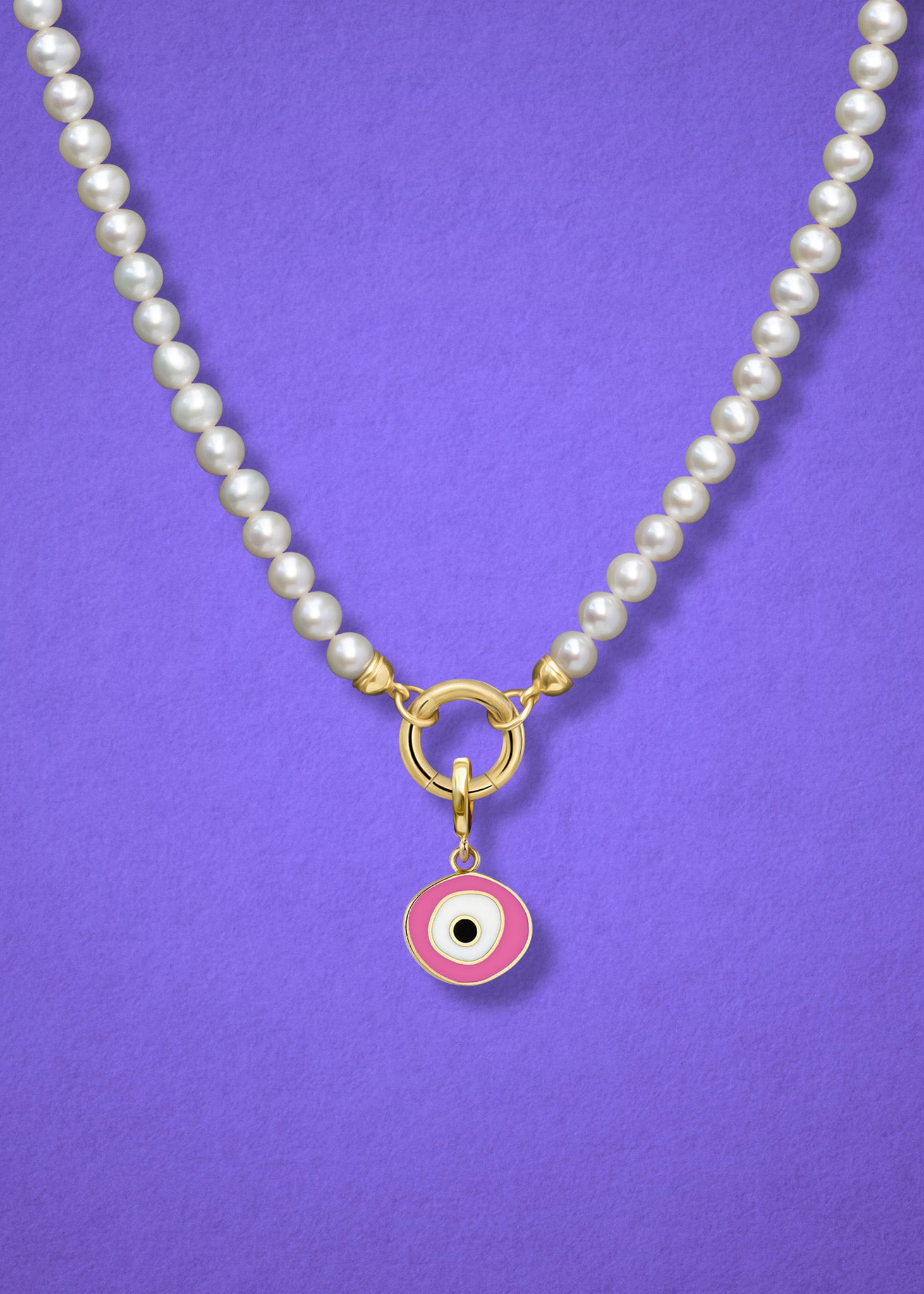 Close-up of the Blush Organic Evil Eye Charm with pink, white, and black enamel on the Lustrous Pearl necklace, shown on a lilac background.
