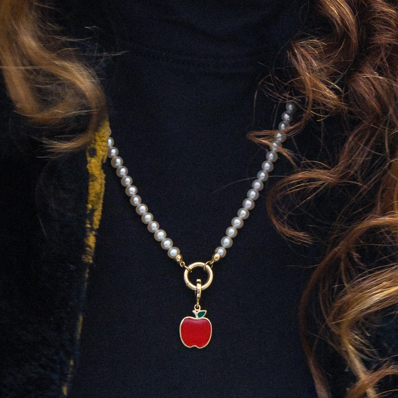 Close-up lifestyle photo of the Lustrous Pearl Golden Apple Charm Necklace in 14K solid gold with a red enamel golden apple of NYC charm, styled on a model wearing a black turtleneck, and a vintage yellow-gold patterned jacket."