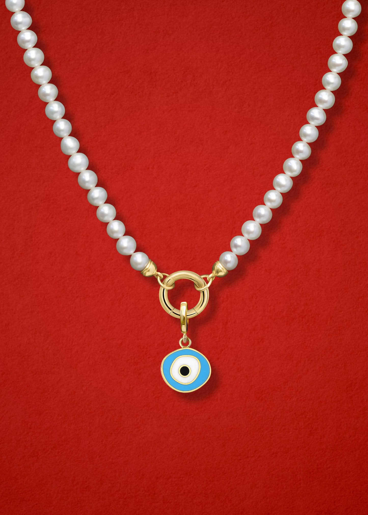 Close-up of the Sky Organic Evil Eye Charm with sky blue, white, and black enamel on the Lustrous Pearl necklace, shown on a red background.