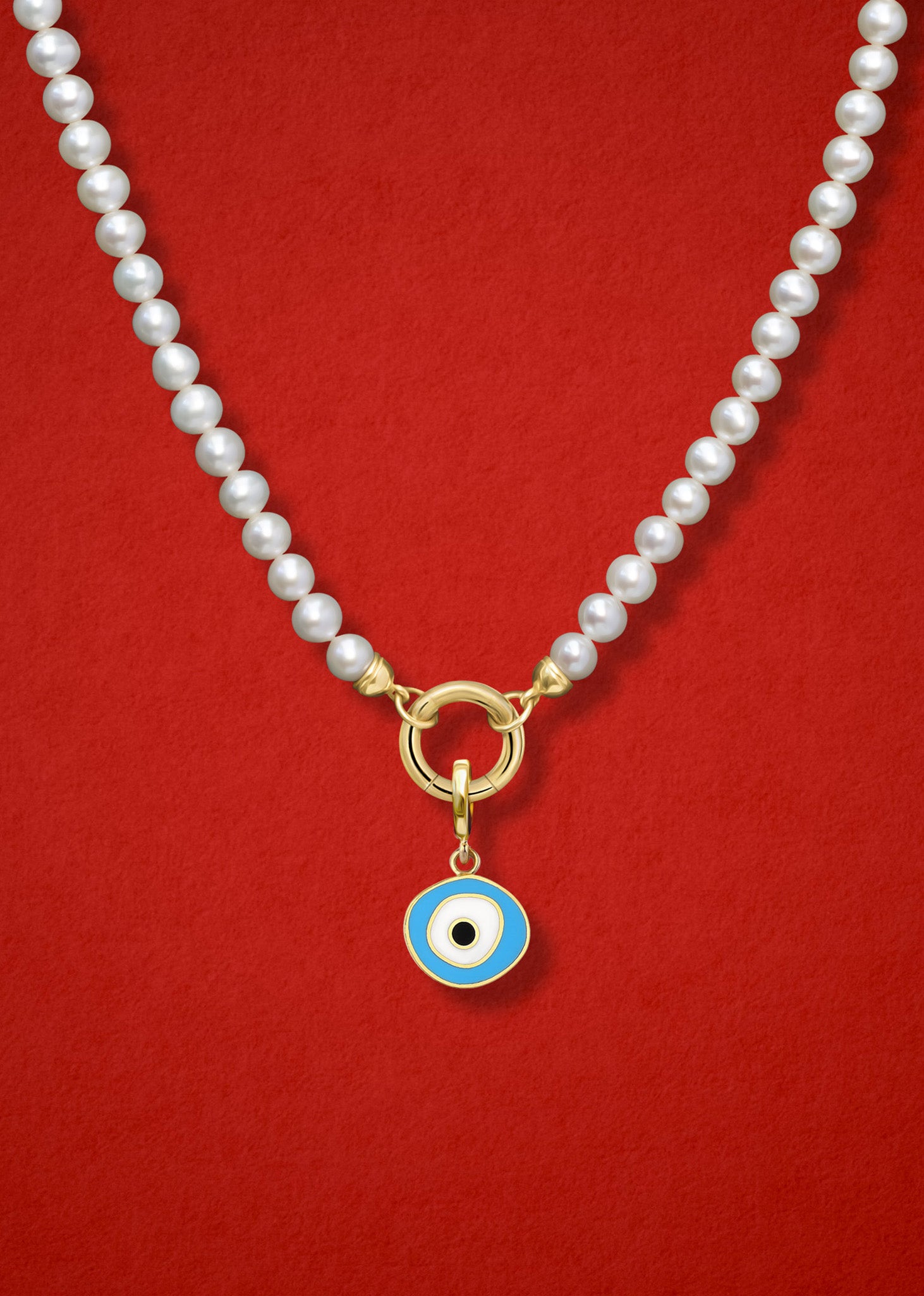 Close-up of the Sky Organic Evil Eye Charm with sky blue, white, and black enamel on the Lustrous Pearl necklace, shown on a red background.