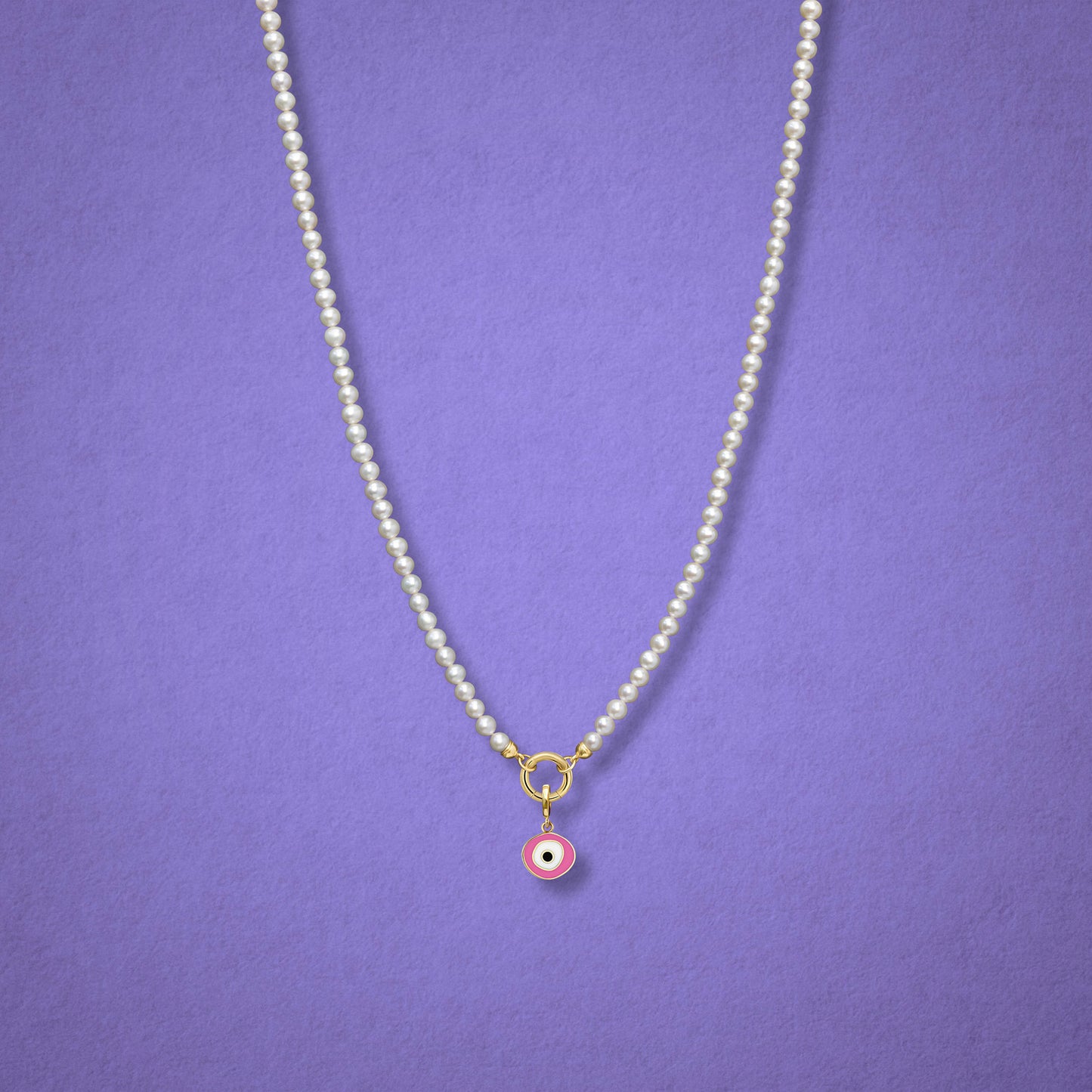 Blush Organic Evil Eye Charm with pink, white, and black enamel shown on the Lustrous Pearl necklace for scale on a lilac background.