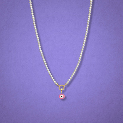 Blush Organic Evil Eye Charm with pink, white, and black enamel shown on the Lustrous Pearl necklace for scale on a lilac background.