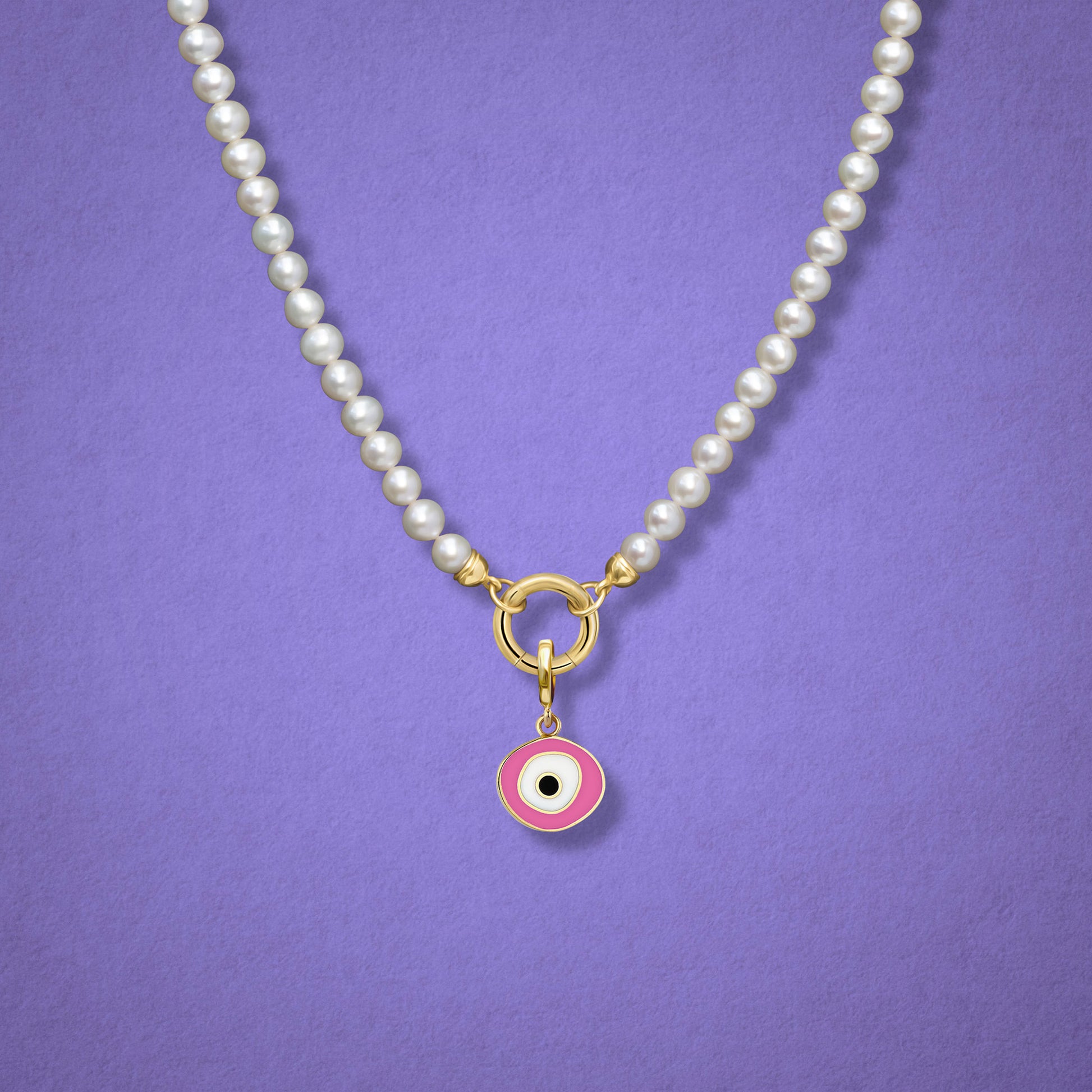Close-up of the Blush Organic Evil Eye Charm with pink, white, and black enamel on the Lustrous Pearl necklace, shown on a lilac background.