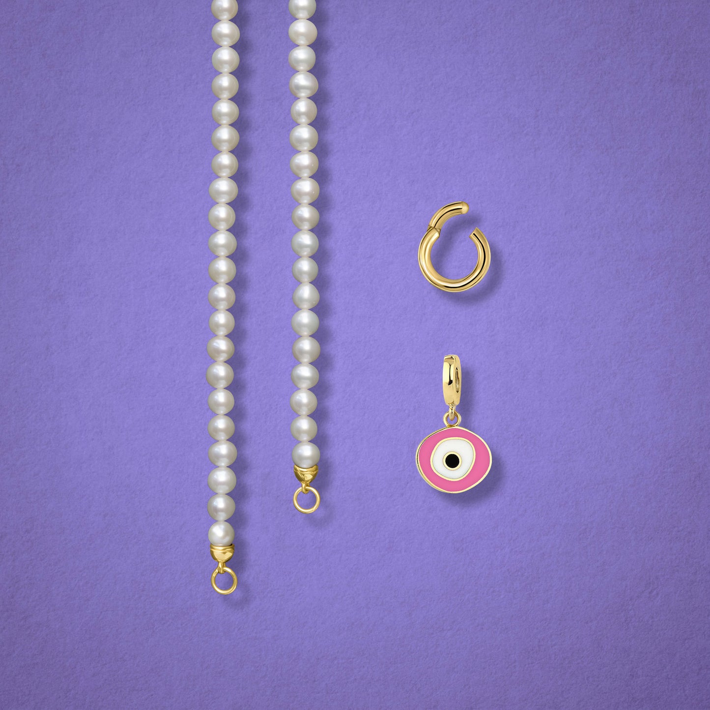 Close-up of the Blush Organic Evil Eye clip-on charm, Golden Link Charm Holder, and the Lustrous Pearl necklace displayed side by side on a lilac background.