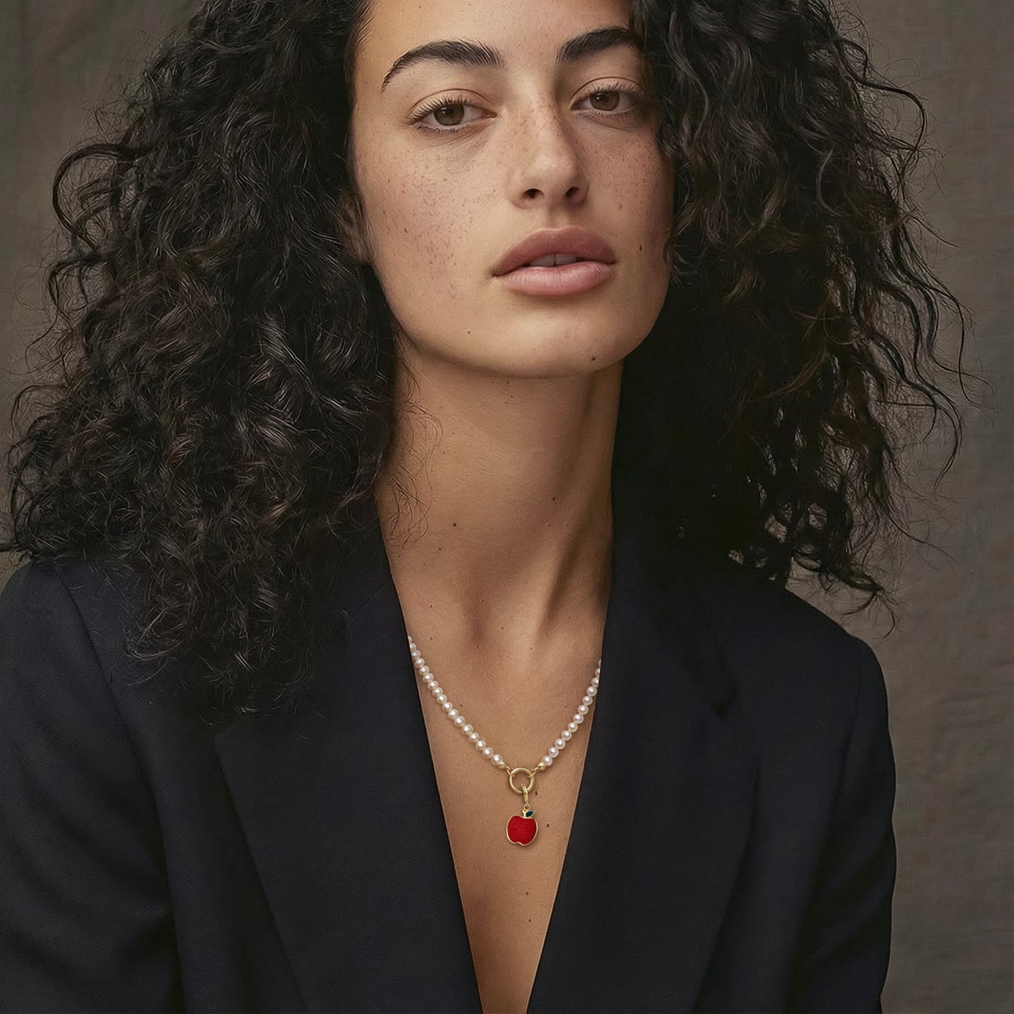 Model wearing the Lustrous Pearl Golden Apple of NYC Charm Necklace, featuring a 20-inch strand of 3.5mm freshwater pearls, 14K solid gold clips, an 11mm outer diameter, a 1.5mm thick 14K solid gold charm holder, and a green and red enamel apple charm crafted in 14K solid gold.