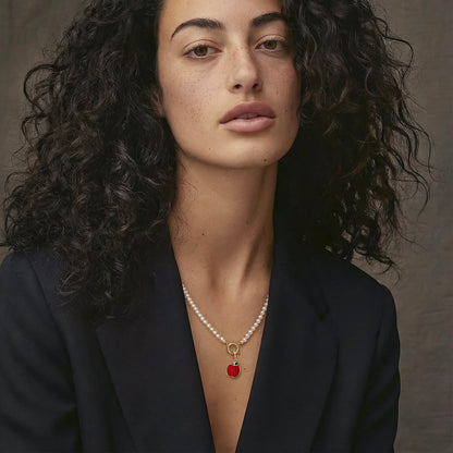 Model wearing the Lustrous Pearl Golden Apple of NYC Charm Necklace, featuring a 20-inch strand of 3.5mm freshwater pearls, 14K solid gold clips, an 11mm outer diameter, a 1.5mm thick 14K solid gold charm holder, and a green and red enamel apple charm crafted in 14K solid gold.