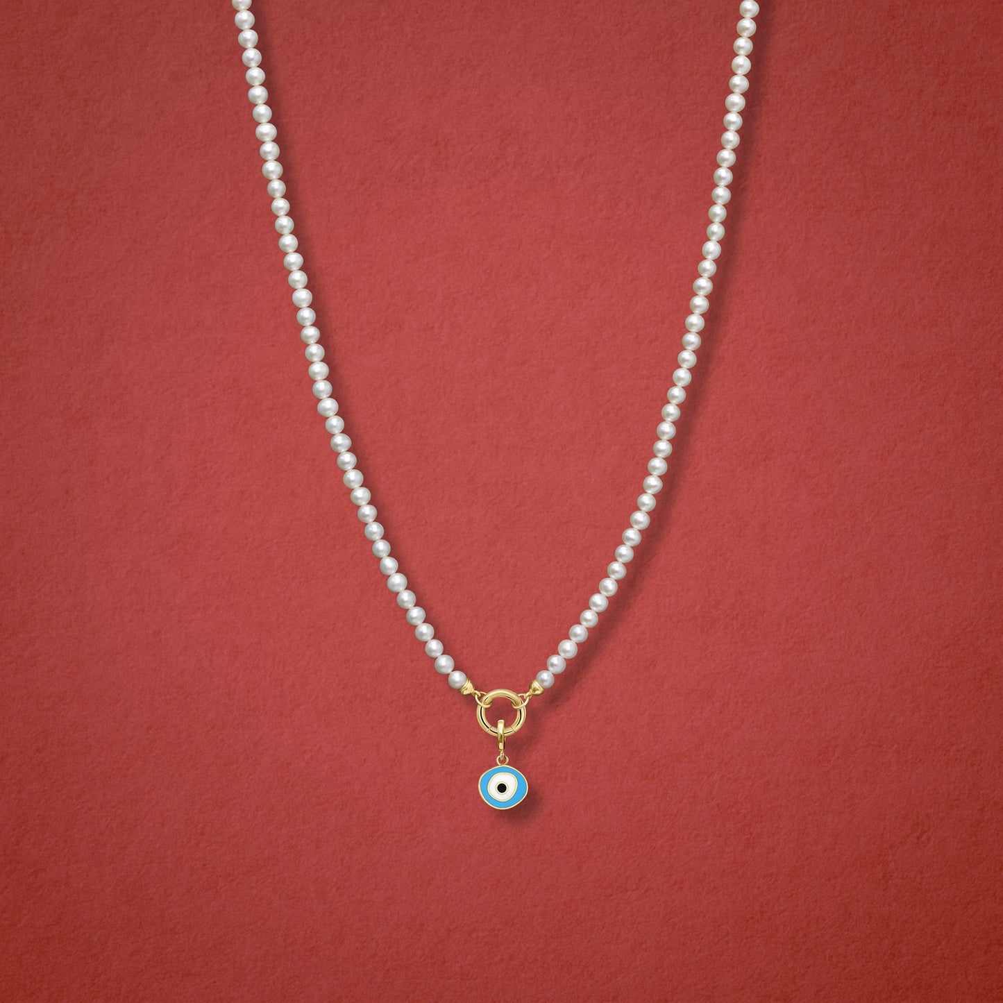 Sky Organic Evil Eye Charm, featuring blue, white, and black enamel, shown on the Lustrous Pearl necklace for scale against a red background.