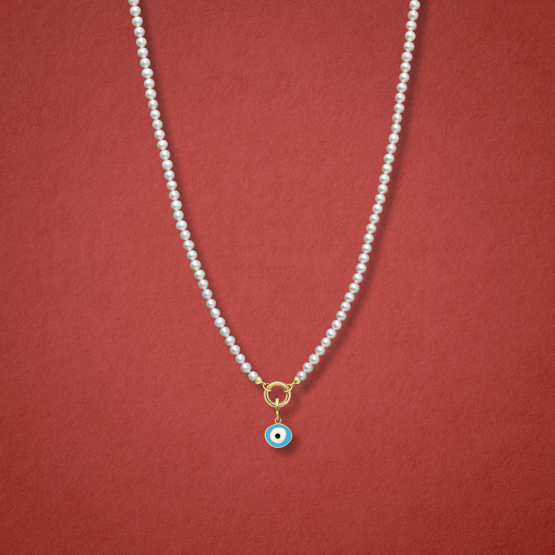 Sky Organic Evil Eye Charm, featuring blue, white, and black enamel, shown on the Lustrous Pearl necklace for scale against a red background.