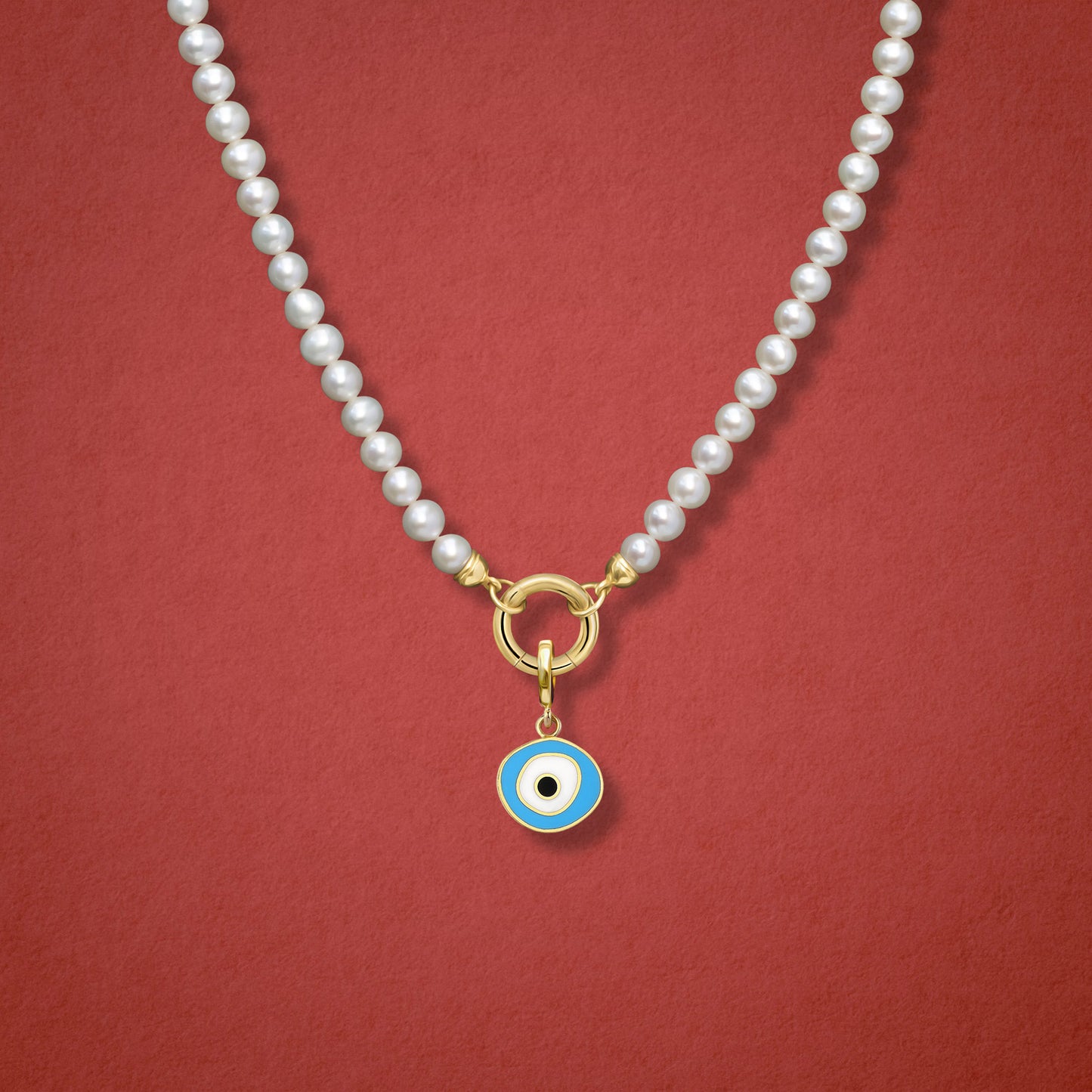 Close-up of the Sky Organic Evil Eye Charm with blue, white, and black enamel on the Lustrous Pearl necklace, shown on a red background.