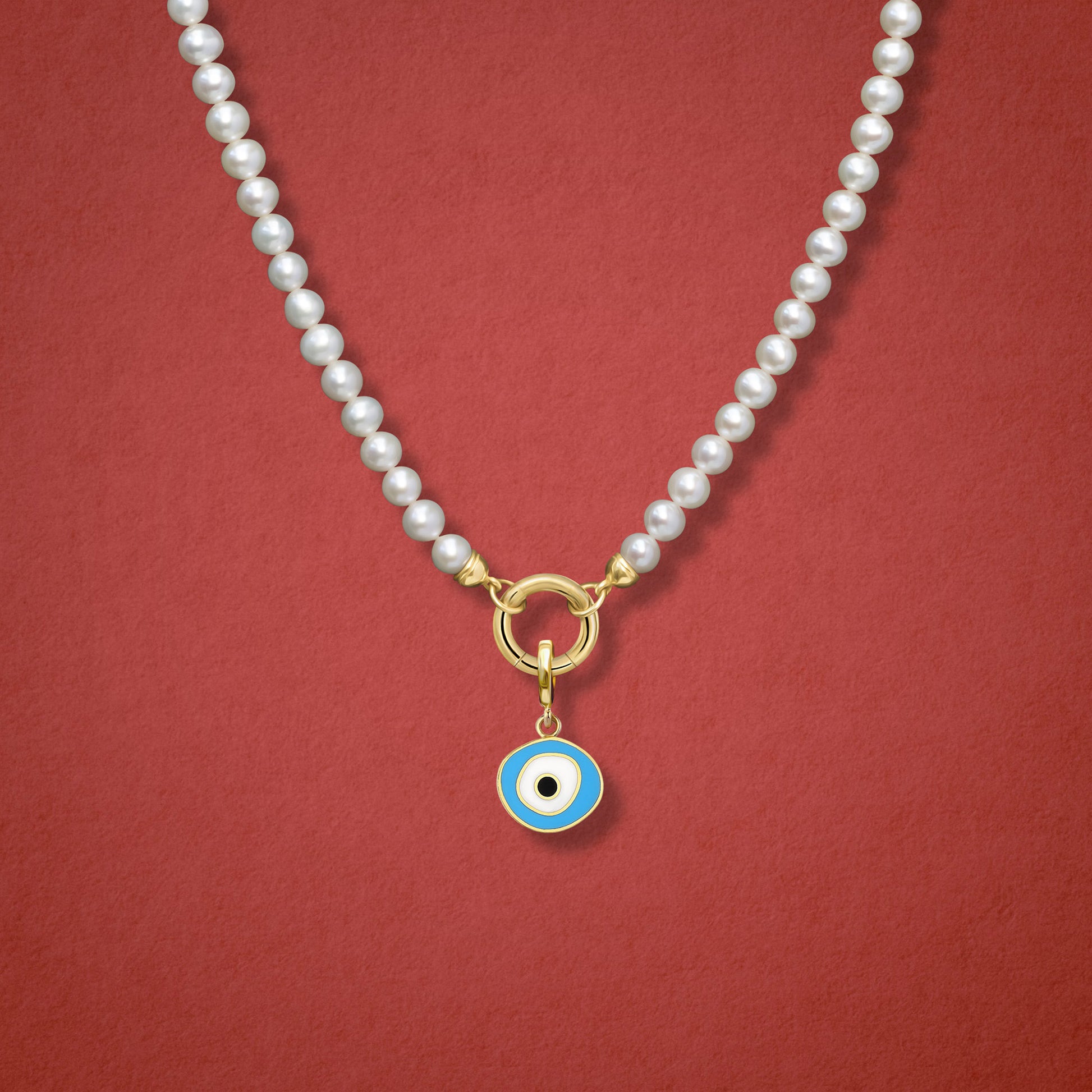 Close-up of the Sky Organic Evil Eye Charm with blue, white, and black enamel on the Lustrous Pearl necklace, shown on a red background.