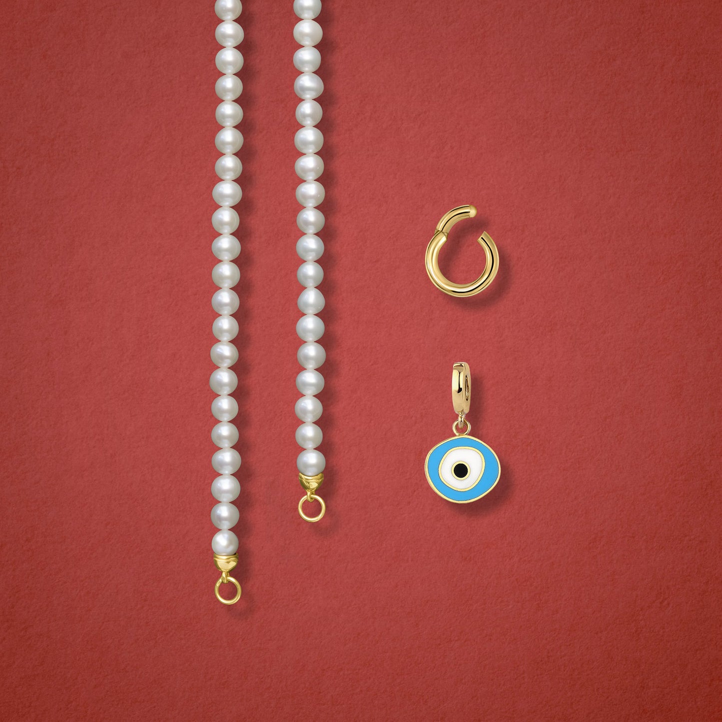 Close-up of the Sky Organic Evil Eye clip-on charm, Golden Link Charm Holder, and the Lustrous Pearl necklace displayed side by side on a red background.