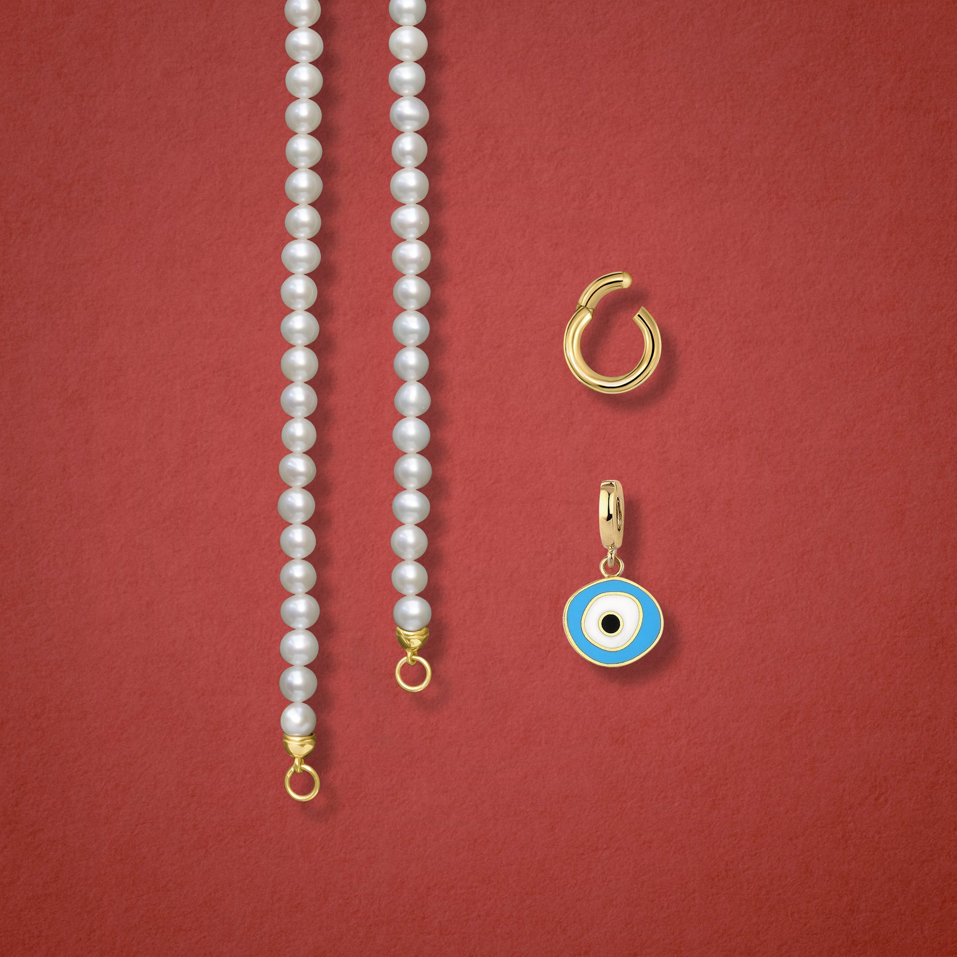 Close-up of the Sky Organic Evil Eye clip-on charm, Golden Link Charm Holder, and the Lustrous Pearl necklace displayed side by side on a red background.