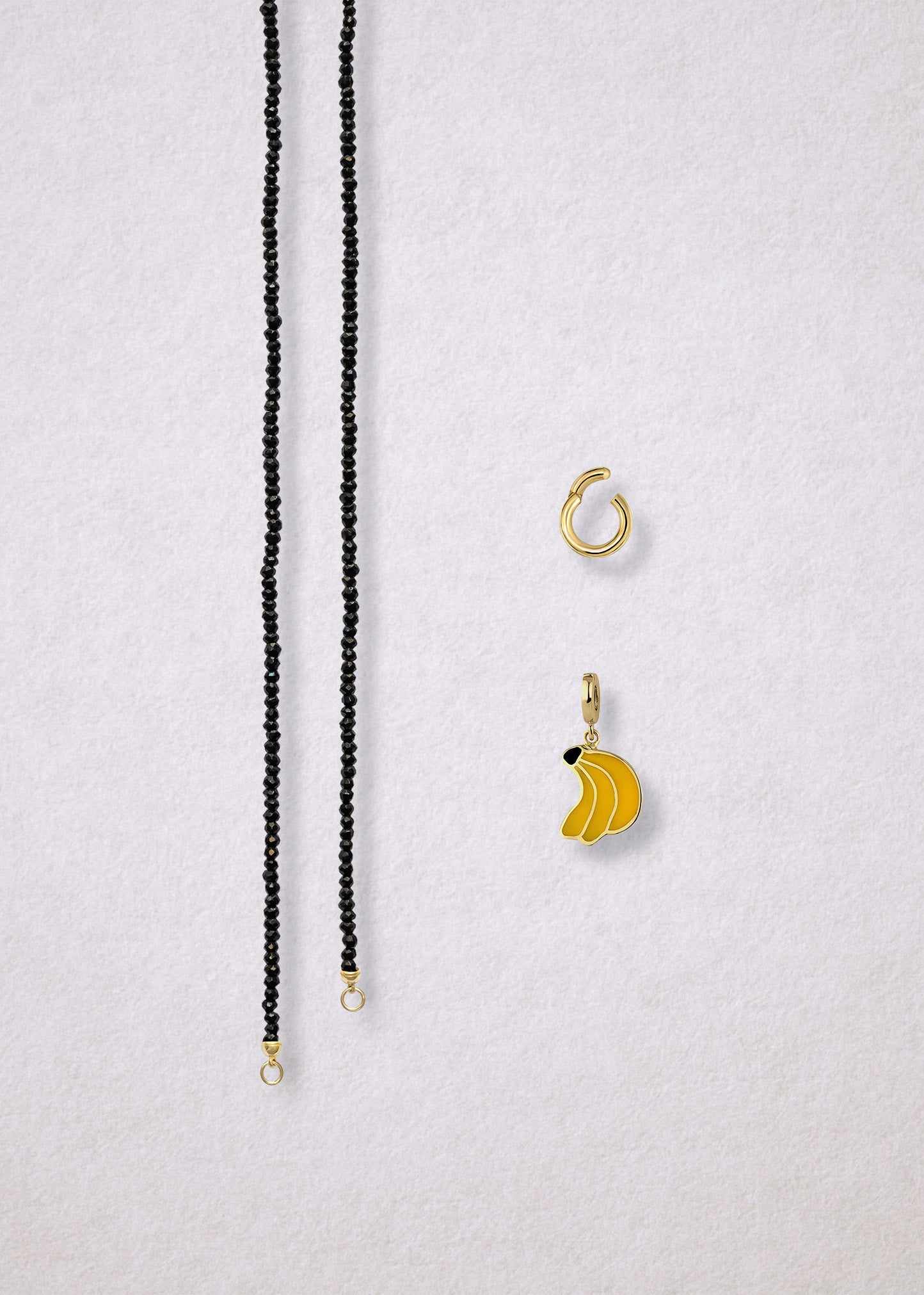 Separated components of the Noir Elegance Banana Bliss Charm Necklace, including faceted black spinel necklace, golden link charm holder, and banana bliss charm with yellow enamel by whywhy NYC.