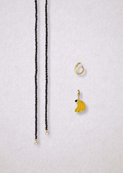 Separated components of the Noir Elegance Banana Bliss Charm Necklace, including faceted black spinel necklace, golden link charm holder, and banana bliss charm with yellow enamel by whywhy NYC.