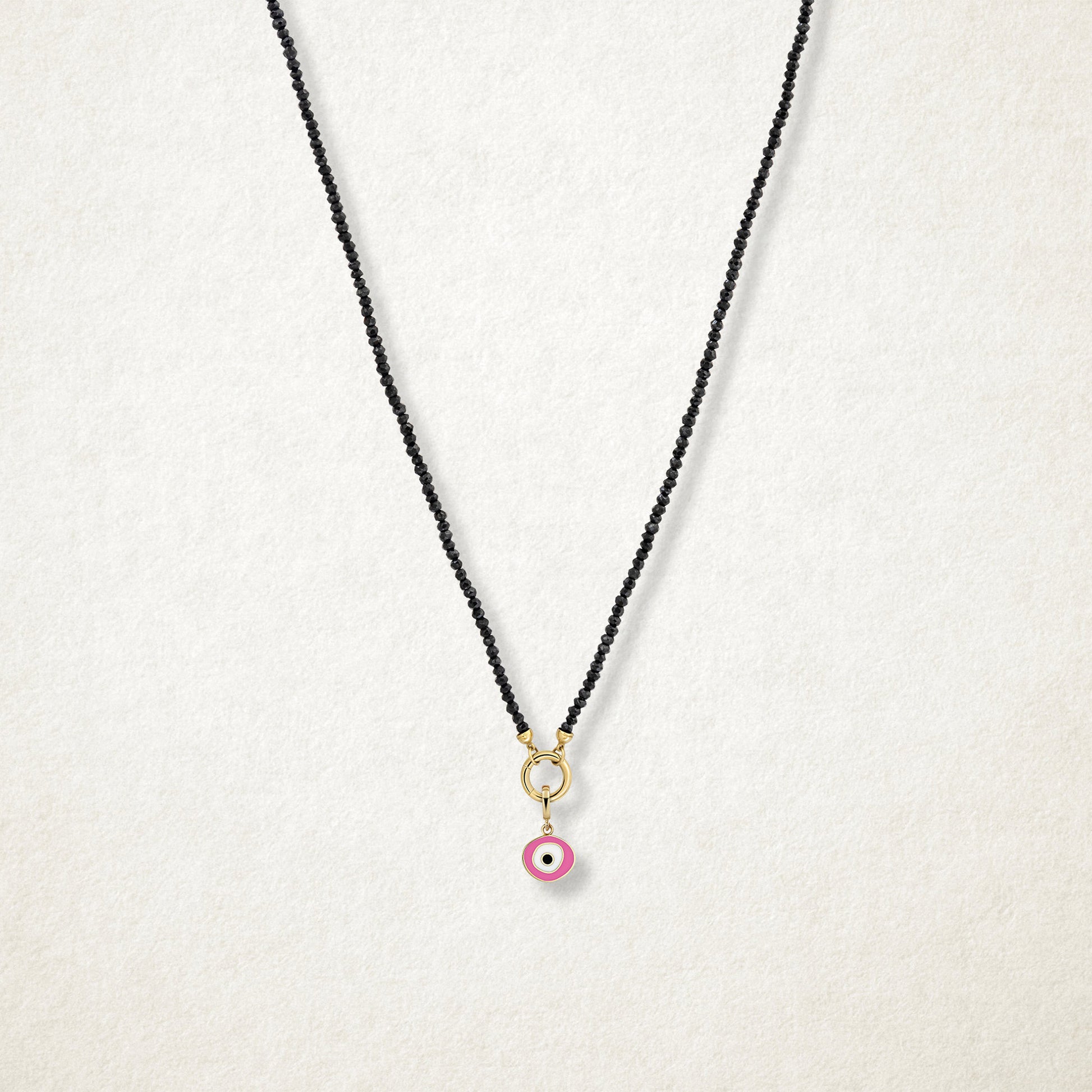 Blush Organic Evil Eye Charm with pink, white, and black enamel shown on the Noir Elegance black spinel necklace for scale on a soft light background.