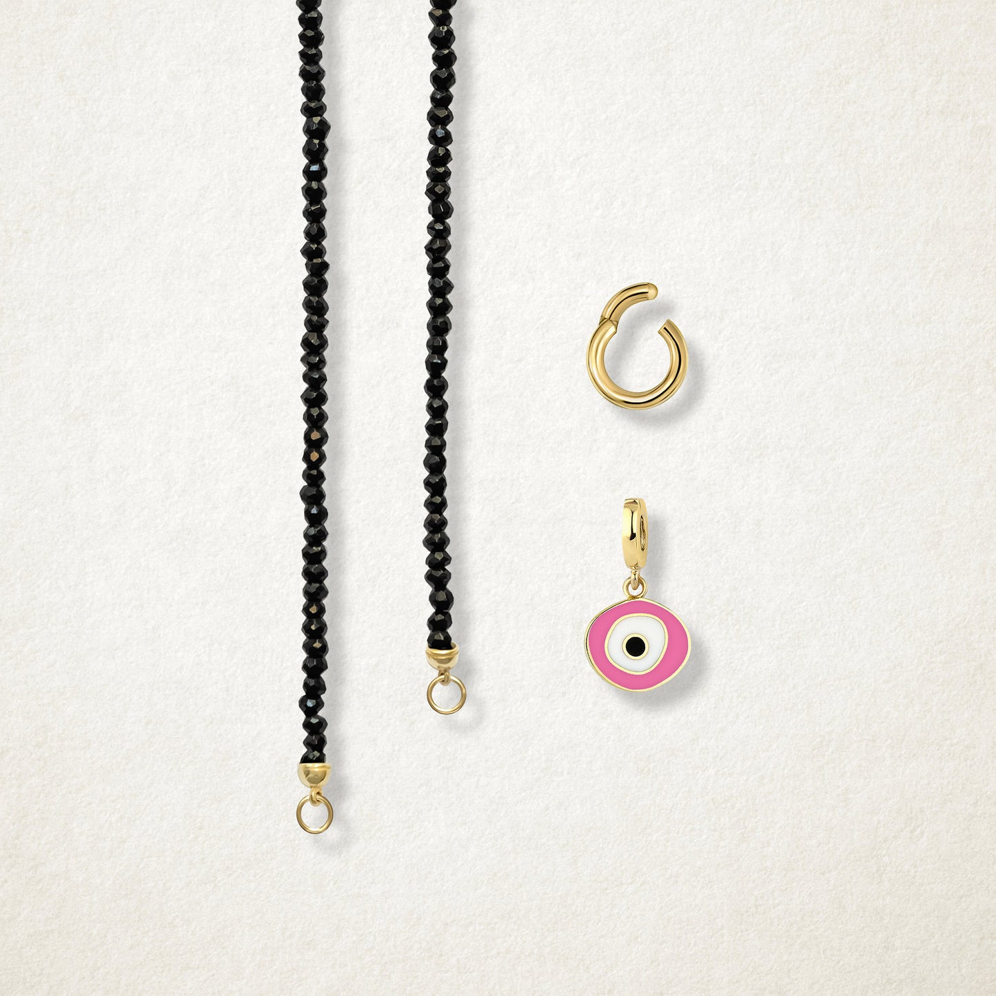 Close-up of the separated Noir Elegance Blush Organic Charm Necklace components: black spinel necklace, golden link charm holder, and pink nazar charm with pink, white and black enamel by whywhy NYC.