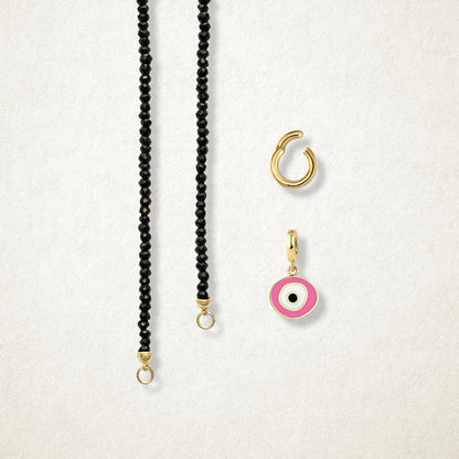 Close-up of the separated Noir Elegance Blush Organic Charm Necklace components: black spinel necklace, golden link charm holder, and pink nazar charm with pink, white and black enamel by whywhy NYC.