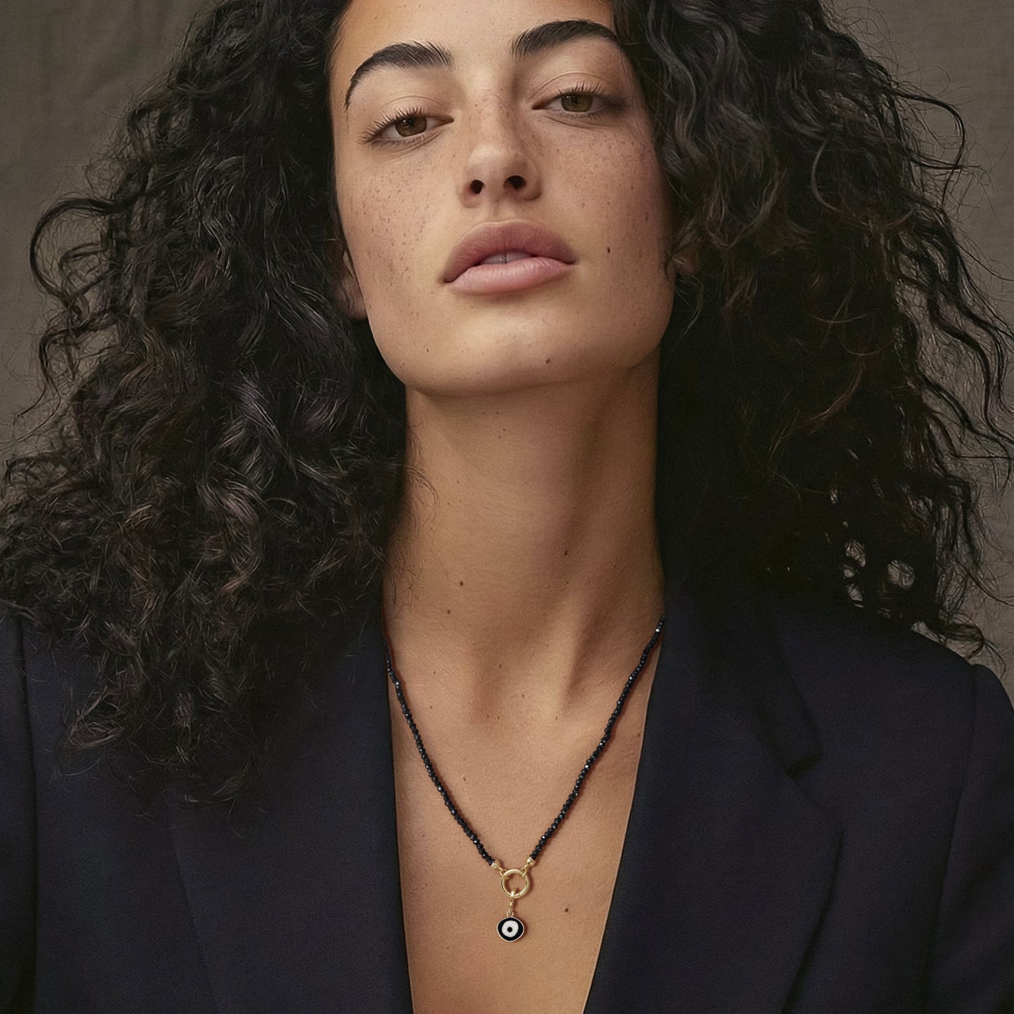 Model wearing the Noir Elegance Obsidian Organic Evil Eye Charm Necklace, featuring 2mm black spinel beads, 14K solid gold clips, an 11mm outer diameter, 1.5mm thick 14K solid gold charm holder, and a black and white enamel evil eye charm crafted in 14K solid gold.