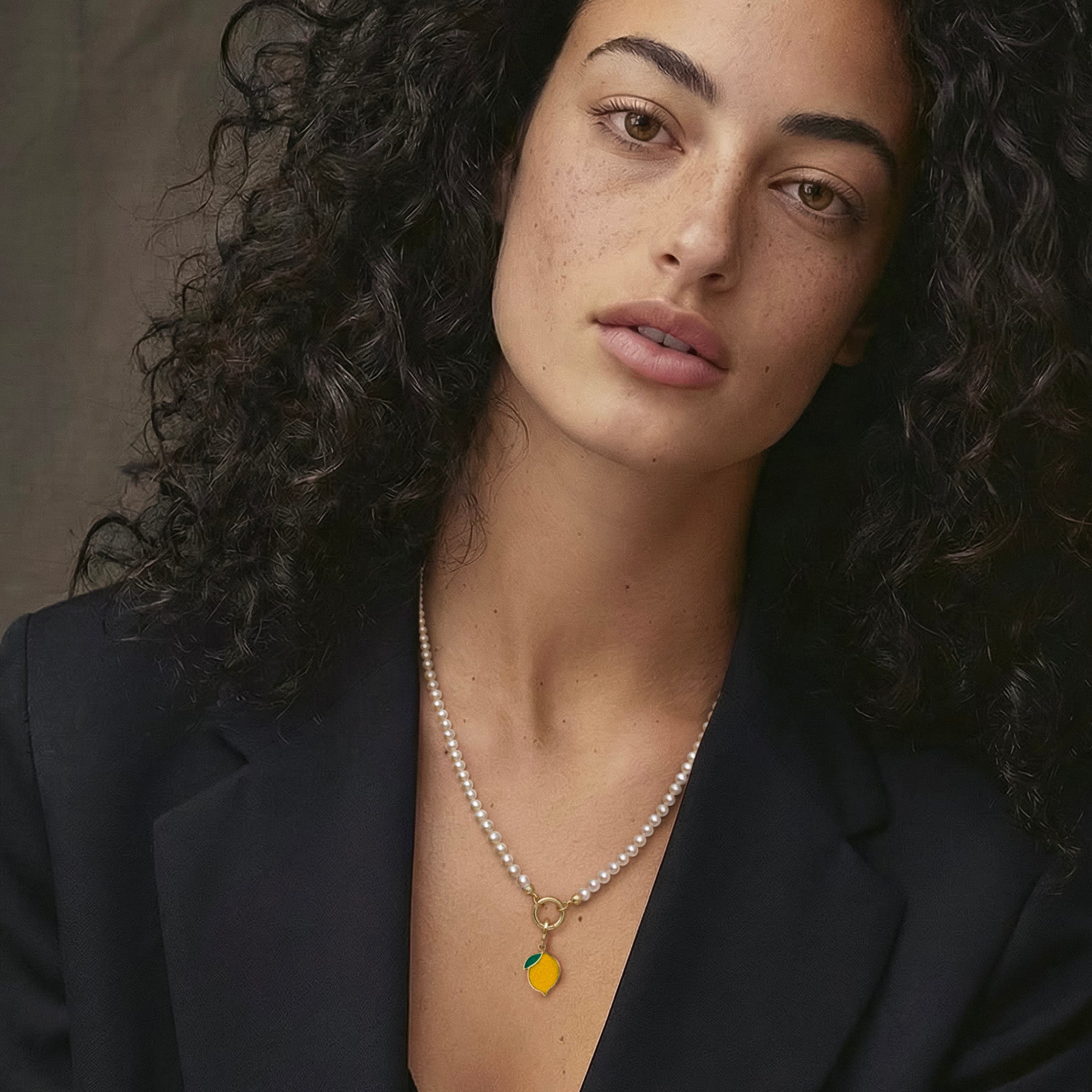 Model wearing the Lustrous Pearl Zesty Lemon Charm Necklace, featuring a 20-inch strand of 3.5mm freshwater pearls, 14K solid gold clips, an 11mm outer diameter, a 1.5mm thick 14K solid gold charm holder, and a green and yellow enamel lemon charm crafted in 14K solid gold.